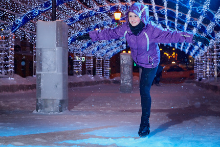 Girl skating on the ice arena full of light on the city square in winter evening. Photo shoot with a teenager female athlete with blue lights on a snowy winter nightの写真素材