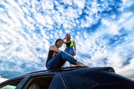Two girls on the roof of a big black car and white clouds above her in a summer eveningの写真素材