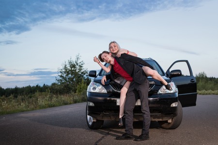 Guy carrying the girl on the road near the car in a summer evening. Funny photoshoot