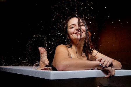 Young pretty brunette woman in bath with water and splashes and drops. Model during an unusual photo shoot in a dark roomの写真素材