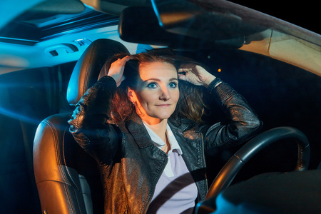 Fashionable young woman in a black leather jacket in a car in the black night and colour light...