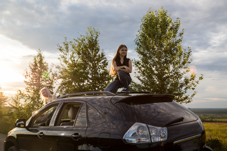 Girl on the roof of a big black car and white clouds above her in a summer eveningの写真素材