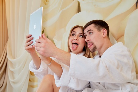 Couple of tourists resting in beautiful hotel room on vacations. Honemoon travelers in white bathrobe in the wedding room. Boy and a girl doing a selfie for tabletの写真素材