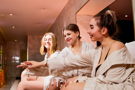 Three cute girls in bath robes on wooden chairs in the indoor pool. Female friends on vacation at the Spa on the sun bedの写真素材