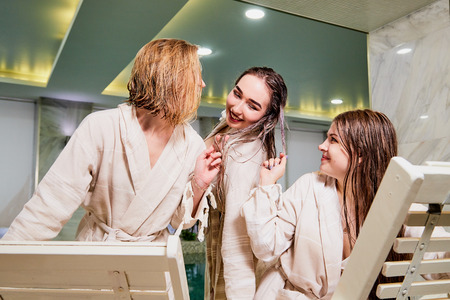 Three cute girls in bath robes on wooden chairs in the indoor pool. Female friends on vacation at the Spa on the sun bedの写真素材