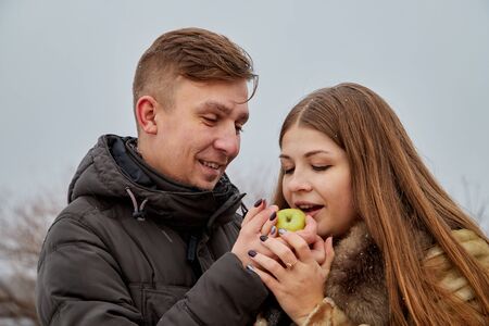 Romantic couple in love on autumn or winter walk near lake. The guy treats the girl with an Apple