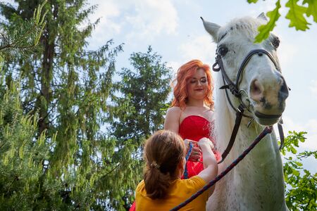 Kirov, Russia - May 30, 2019: Photo shoot girl with red hair and red dress with white horse and trainer groom helping her. Unusual posing with an animal in the summer dayのeditorial素材