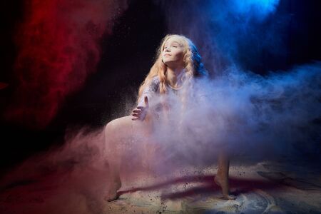 Beautiful teen girl with long blonde curly hair in a dark room with colored lights and clouds of flour. Sports teenager young model during a photoshoot with flying flour and color lightの写真素材