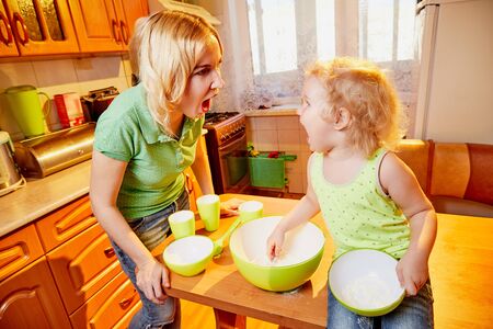 Mother and her three years old blonde daughter are arguing and swearing in the kitchen during cooking. Funny photoshoot of a little girl and an adult womanの写真素材