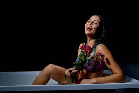 Young pretty brunette woman in bath with water and flowers. Model during an unusual photo shoot in a dark roomの写真素材