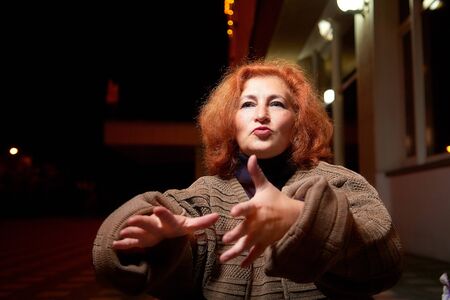 Emotional middle-aged woman in a thick gray sweater on a city street at night. Elderly female...