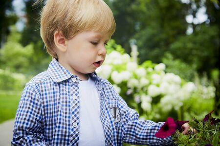 Cute baby blonde boy in a park or a garden near flowers in a summer, spring or autumn day. Outdoor...