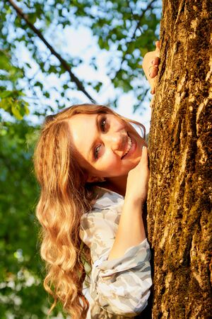 Beautirul girl with curly blonde hair near a tree in the park with sunny weather in a summer, spring or autumn dayの写真素材
