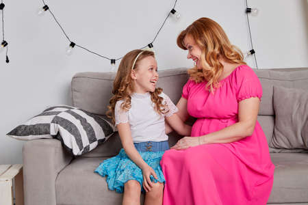 Happy pregnant mother and daughter in bright summer dresses having conversation and smiles together....
