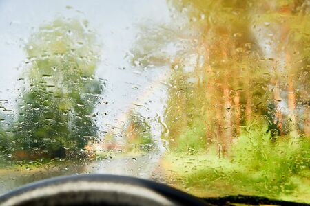 View from car cabin on wet asphalt road, trees and rainbow during rain and drops on front window....