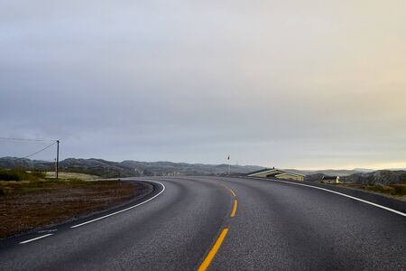 View on grey road, yellow sunrise and dramatic clouds from the car front window in early mourning. Driving car during sunshine in the tundra in Norway. Background of nature landscape and grey roadの写真素材