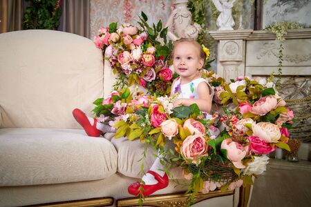 Cute blonde small girl sitting on sofa in living room with beautiful flowers. Photoshoot in studio