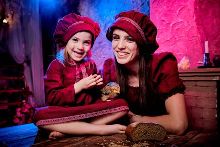 Easter and family. Mother and daughter in maroon or red dresses of village shepherdesses and small chicken in rural wooden interior in studio. Colour beautiful light background of two female relativesの写真素材