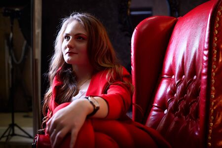Girl in red costume sitting in red chair in the dark room. Model posing during fashion photoshoot