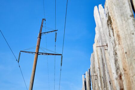 Electric power lines and wires with blue sky and wooden fence. Support of power lines in a village...