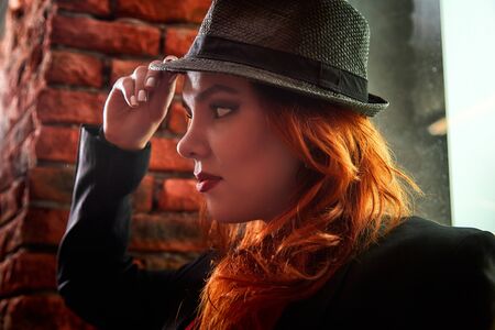 Beautiful young woman with redhair, black hat and red clothes being near red brick wall indoors