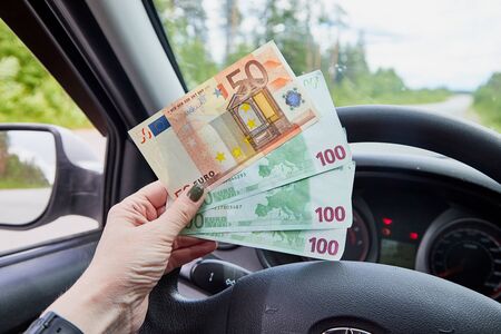 Euro banknotes of various denominations in a woman hand inside of a car and road with nature outside