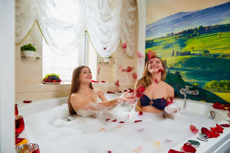 Kirov, Russia - February 27, 2019: Two girls have fun in modern bath full of white foam and red flower petals, with green pictures on the wall and window. Female friends having rest and relaxationのeditorial素材