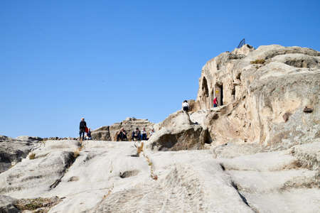Uplistsikhe, Georgia - October 22, 2020: Tourists in a mountain reserve near ancient old city with rocks, residential and religious caves and blue sky on Sunny summer day. Uplistsikhe city in Georgiaのeditorial素材