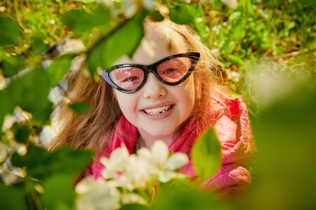Pretty cute little girl with long hair and in pink glasses posing near a blooming apple tree with white flowers in a summer or spring parkの写真素材