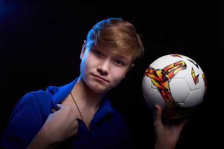 Low key portrait of a handsome brunette young male teenager in blue shirt with soccer ball in hand. Interesting boy soccer player and dark background with blue light. Sport model posing in photoshootの写真素材