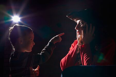 Ugle oman in red sweater and pirate hat with curly hair posing with small boy on black background....