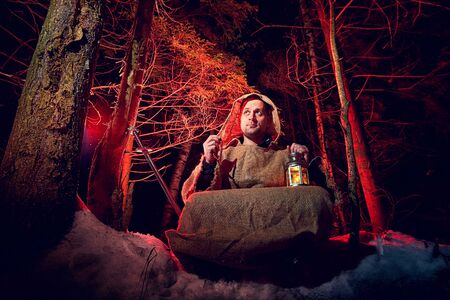 Medieval monk in canvas sackcloth robe praying in dark forest with snow and red light on winter night. Fantasy or fairy tale about wandering monk. Story about the forces of good and evil in the worldの写真素材