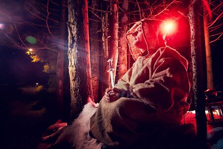 Medieval monk in canvas sackcloth robe praying in dark forest with snow and red light on winter night. Fantasy or fairy tale about wandering monk. Story about the forces of good and evil in the worldの写真素材