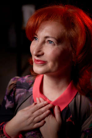 Stylish elderly woman with red hair in vinous dress posing in studio with dark background and red...
