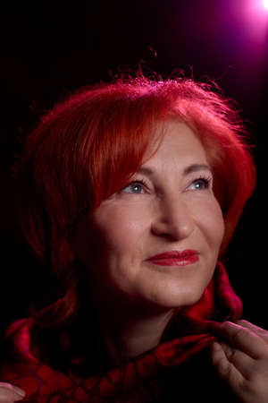 Stylish elderly woman with red hair in vinous dress posing in studio with dark background and red flash and light. Model in photoshootのeditorial素材