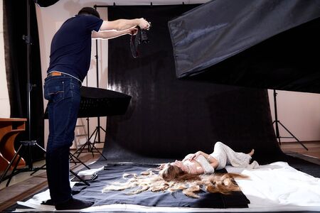 Male photographer taking pictures of model in beautiful shiny dress in photo Studio. Backstage with man and woman during professional shooting indoors. Photoshoot inside of the roomの写真素材