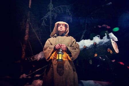 Medieval monk in canvas sackcloth robe with lattern in dark forest with snow and red light on winter night. Fantasy or fairy tale about wandering monk. Story about forces of good and evil in the worldの写真素材