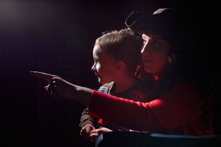 Ugle woman in red sweater and pirate hat with curly hair posing with small boy on black background....