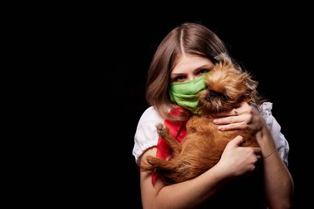 Girl in a protective medical green mask and national ethnic dress holding small dog in her hands. Coronavirus, COVID-19 pandemic concept. Domestic animal allergy, sneezing and coughing near of petsの写真素材