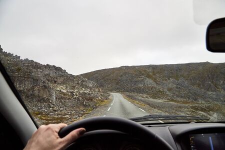 View from car window on the road and pity strange landscape with a mountains, rocks and cloudy sky and hand of woman. Landscape through windscreen in Norway.の写真素材