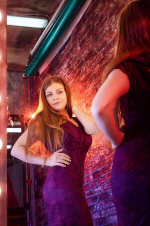 Girl in a burgundy dress in front of a lighted mirror in a dark room. Model posing for a cam during...