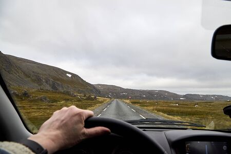 View from car window on the road and pity strange landscape with a mountains, rocks and cloudy sky and hand of woman. Landscape through windscreen in Norway.の写真素材