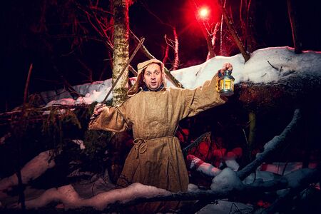 Medieval monk in canvas sackcloth robe with sword and lattern in dark forest and red light of moon on winter night. Fantasy or fairy tale about wandering monk. Concept of forces of good and evilの写真素材