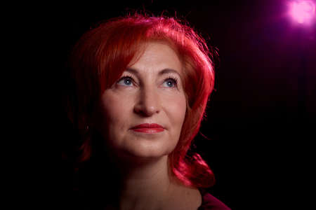 Stylish elderly woman with red hair in vinous dress posing in studio with dark background and red flash and light. Model in photoshootのeditorial素材