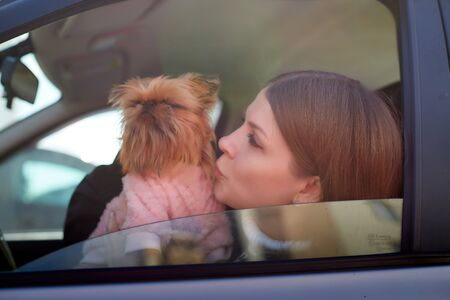 Girl with long hair in a black jacket and a small dog in pink clothes near window of a car. Concept travel with animalの写真素材