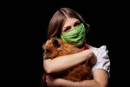 Girl in a protective medical green mask and national ethnic dress holding small dog in her hands. Coronavirus, COVID-19 pandemic concept. Domestic animal allergy, sneezing and coughing near of petsの写真素材