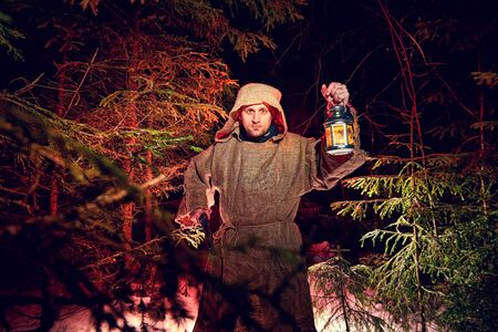 Medieval monk in canvas sackcloth robe with lattern in dark forest with snow and red light on winter...