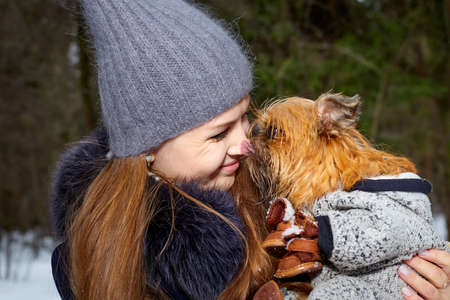 Girl with long hair in black jacket and grey hat posing with a small dog Brussels Griffon in clothes in the forest or park in a winter dayの写真素材