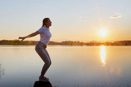 Silhouette of a girl doing sports exercises on the lake and sunset with the sun and rays in the...