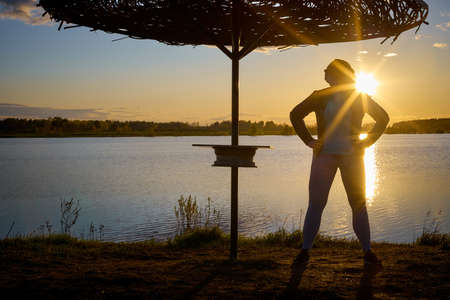 Plump curvy girl with long curly hair near the water of a lake and umbrella at sunset with the sun on the horizon.の写真素材